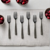 A set of six dinner forks with black heads and geometric patterned handles arranged on a white cloth, surrounded by bowls of berries.